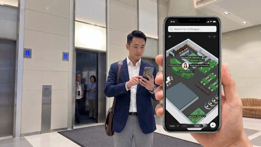エレベーターホールで同僚がスマホを確認。手前の画面は同僚検索で、3Dマップに Sarah J. が Conference Room B にいると表示。